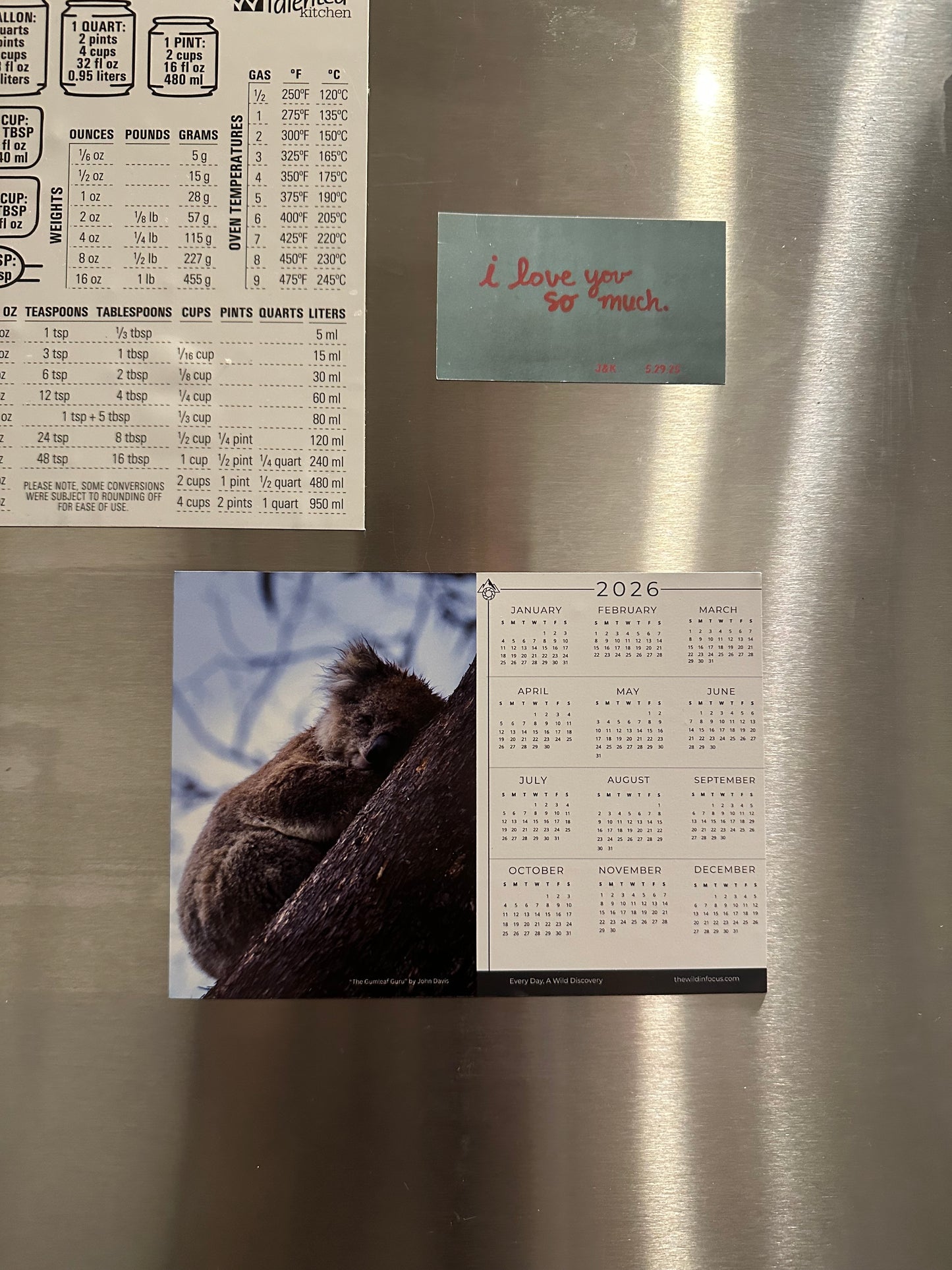 A stainless steel fridge displays The Gumleaf Guru Koala Calendar 2026 - Australian Wildlife Photography Magnet Calendar (Pre-Order), a baking measurement chart, and a blue note with "i love you so much" in red handwriting.