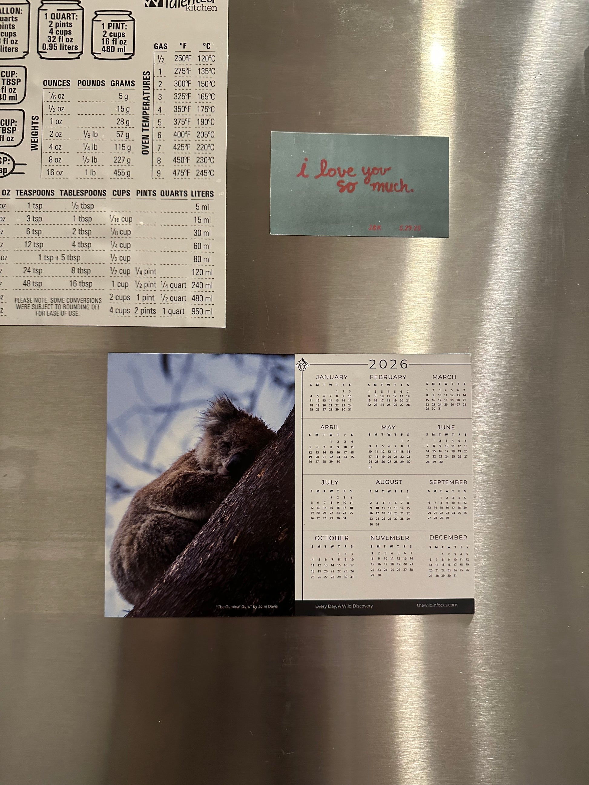 A stainless steel fridge displays The Gumleaf Guru Koala Calendar 2026 - Australian Wildlife Photography Magnet Calendar (Pre-Order), a baking measurement chart, and a blue note with "i love you so much" in red handwriting.
