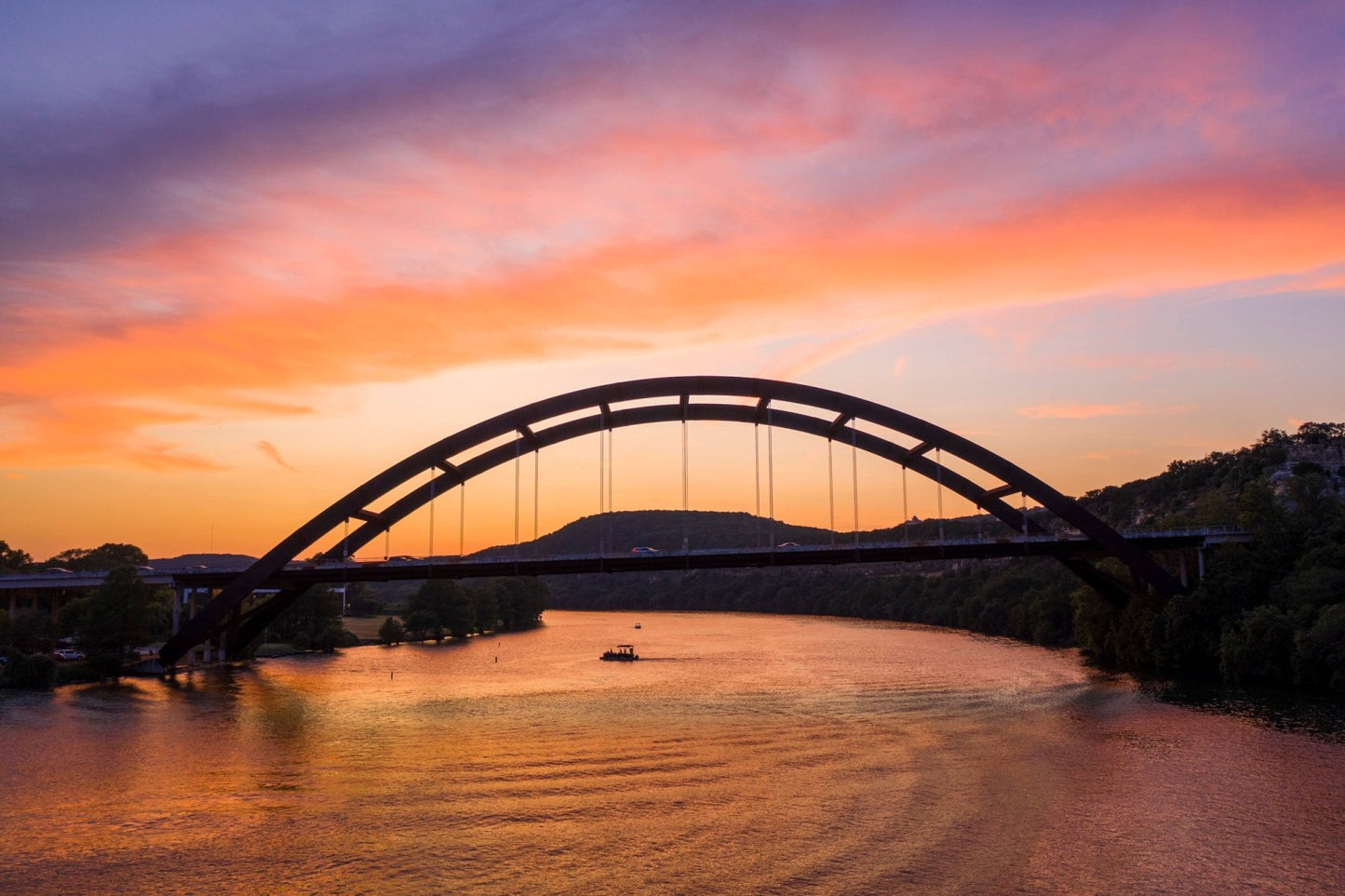 The "Austin Glow" Pennybacker Bridge Sunset Print captures vibrant orange, pink, and purple clouds over the bridge at sunset, with a small boat below and trees along the Texas Hill Country riverbanks. Perfect Austin wall art.