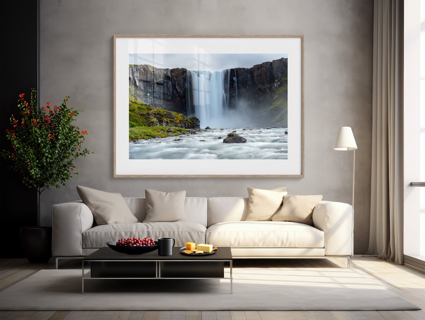A modern living room features a white sofa, beige pillows, a black coffee table, and The Moss Kingdom Falls - Iceland Waterfall Photography Print on the gray wall. A potted plant and floor lamp beside the sofa add warmth to the space.