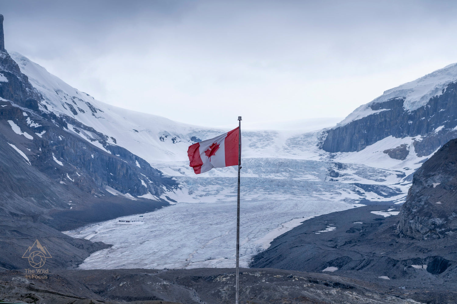 A Canadian flag waves on a pole in front of a glacier nestled between rugged, snow-capped mountains under a cloudy sky.