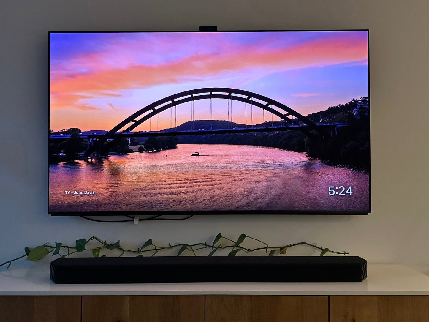 The Austin Glow Digital Bundle displays the Pennybacker Bridge sunset on a Samsung Frame TV. The time shows 5:24. A soundbar sits below on a wooden cabinet, which is decorated with a green leafy vine.