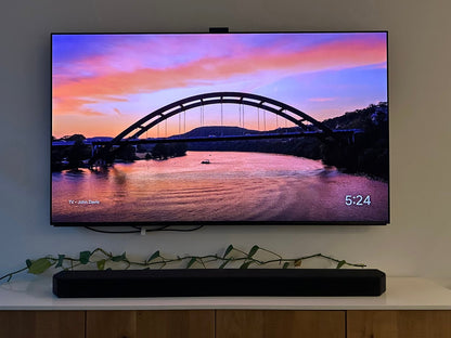 The Austin Glow Digital Bundle displays the Pennybacker Bridge sunset on a Samsung Frame TV. The time shows 5:24. A soundbar sits below on a wooden cabinet, which is decorated with a green leafy vine.