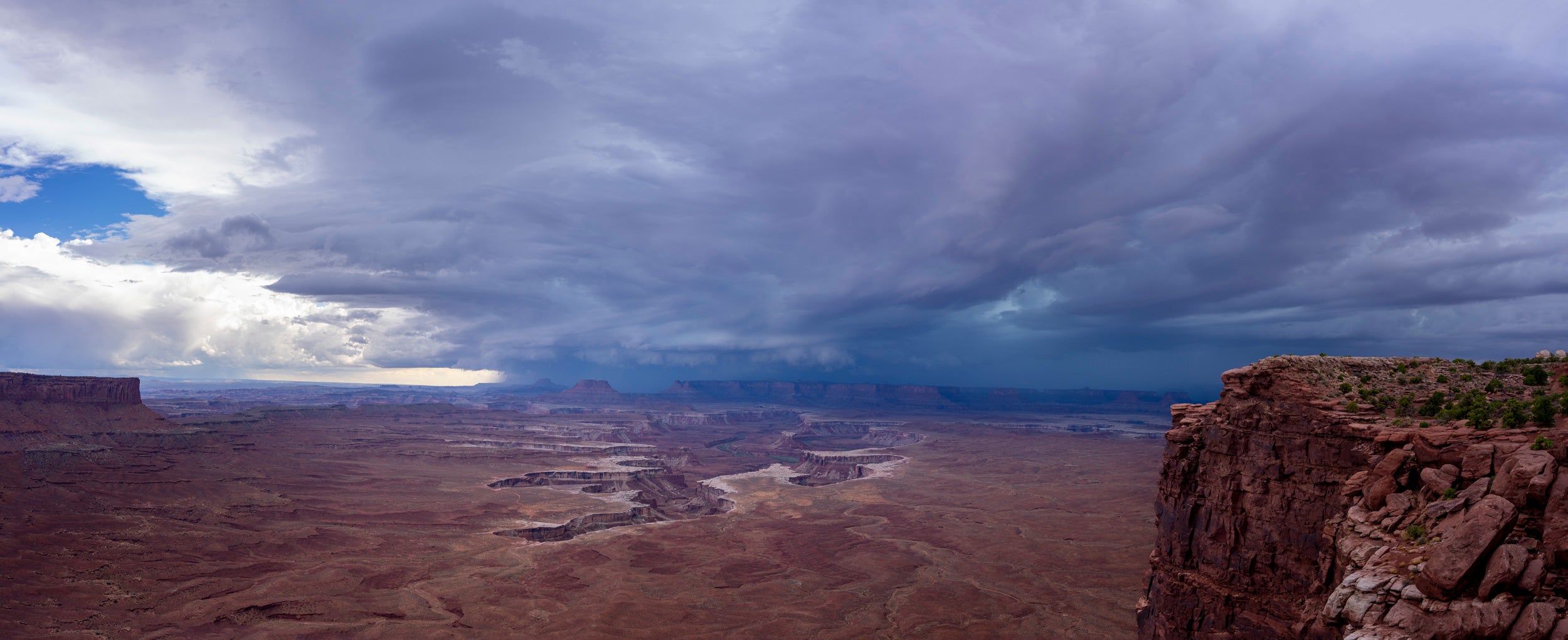 Dramatic storm clouds loom over the vast, rugged red rock canyons and winding river below at Canyonlands National Park, with a steep cliff and sparse vegetation on the right.