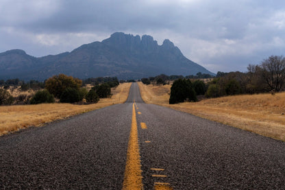 Celestial Highway | West Texas Road Print features a straight road leading to distant mountains under cloudy skies, with dry grass and scattered trees—ideal wall art capturing the striking rural beauty of West Texas.