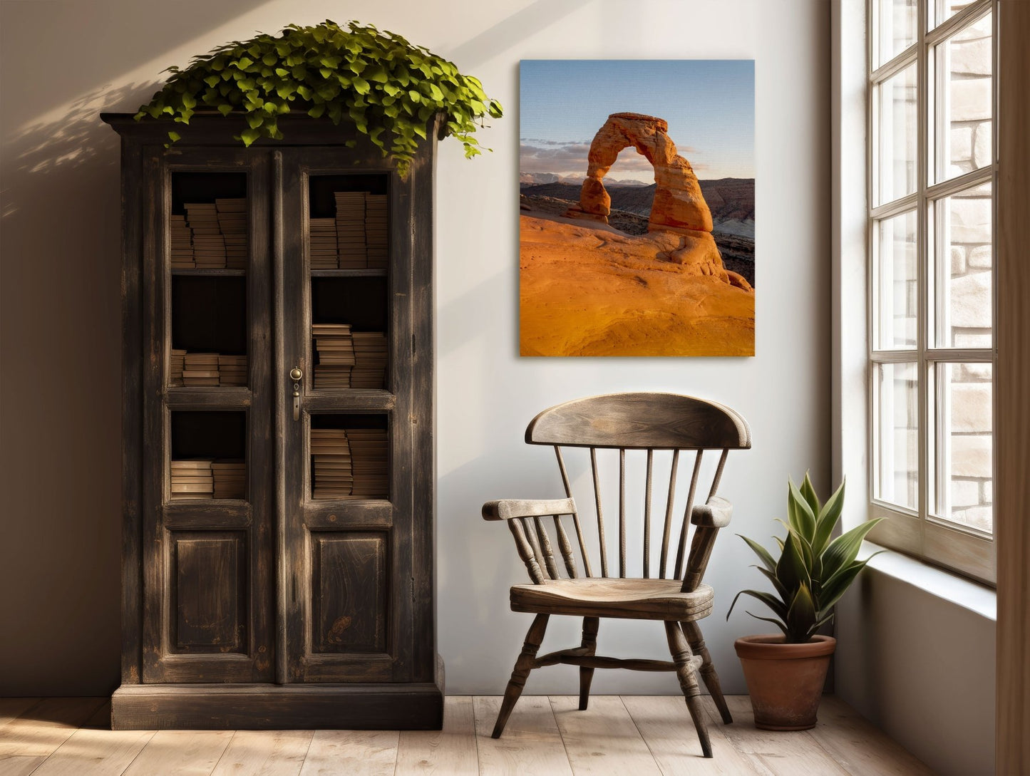 A rustic wooden chair rests by a vintage bookcase filled with books. A potted plant and southwest decor, including the Delicate Light | Vertical Delicate Arch Print, Utah, create a cozy corner by a sunny window.