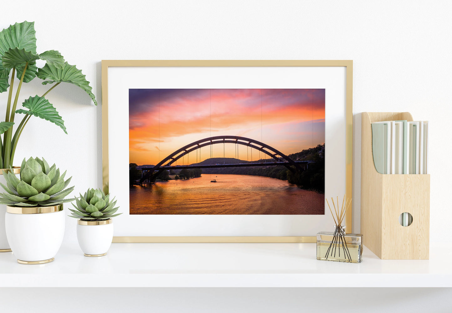 The Austin Glow | Pennybacker Bridge Wall Art decorates a white shelf with potted succulents, a reed diffuser, and a wooden file holder filled with books—a neatly arranged, bright scene.