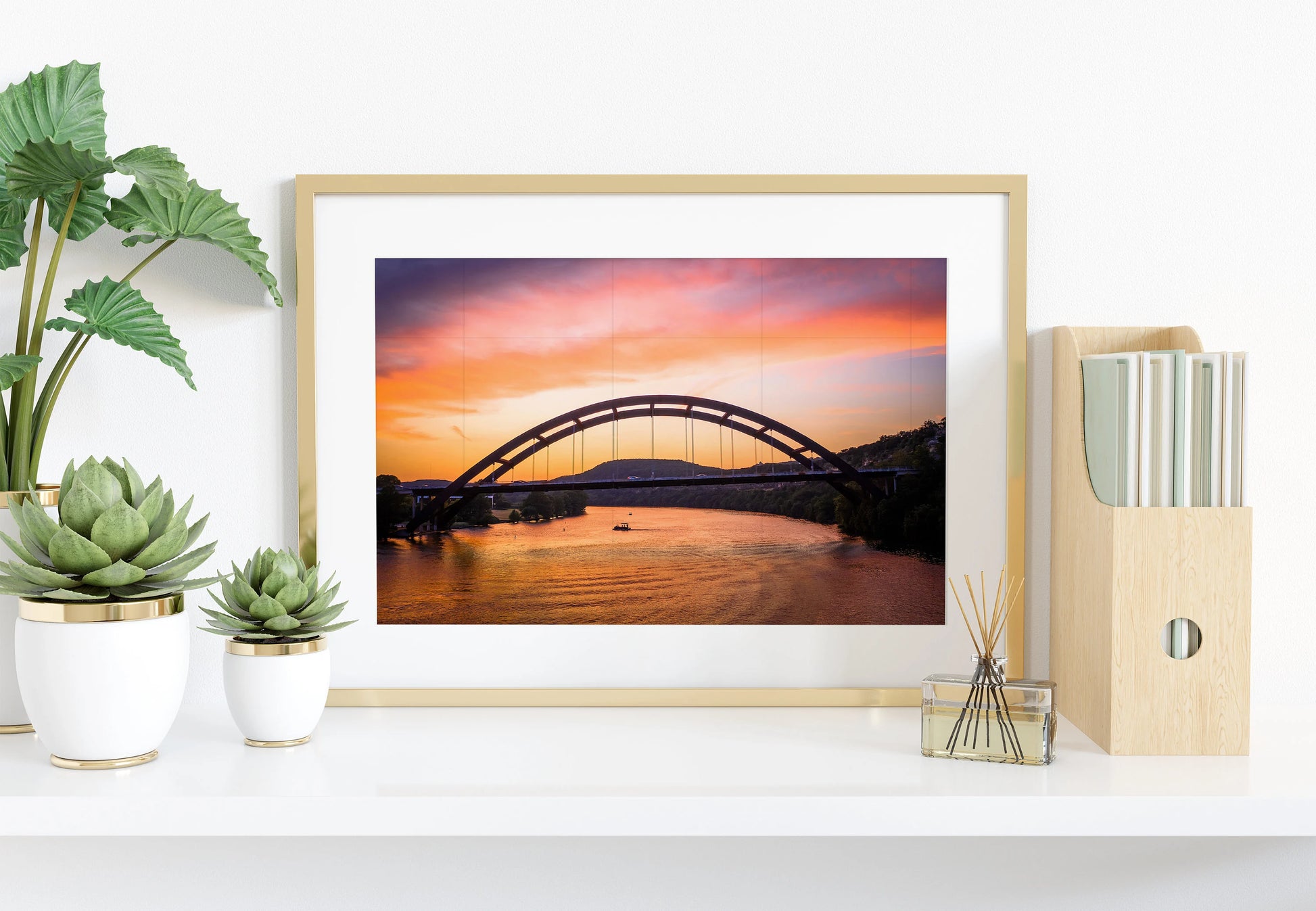 The Austin Glow | Pennybacker Bridge Wall Art decorates a white shelf with potted succulents, a reed diffuser, and a wooden file holder filled with books—a neatly arranged, bright scene.