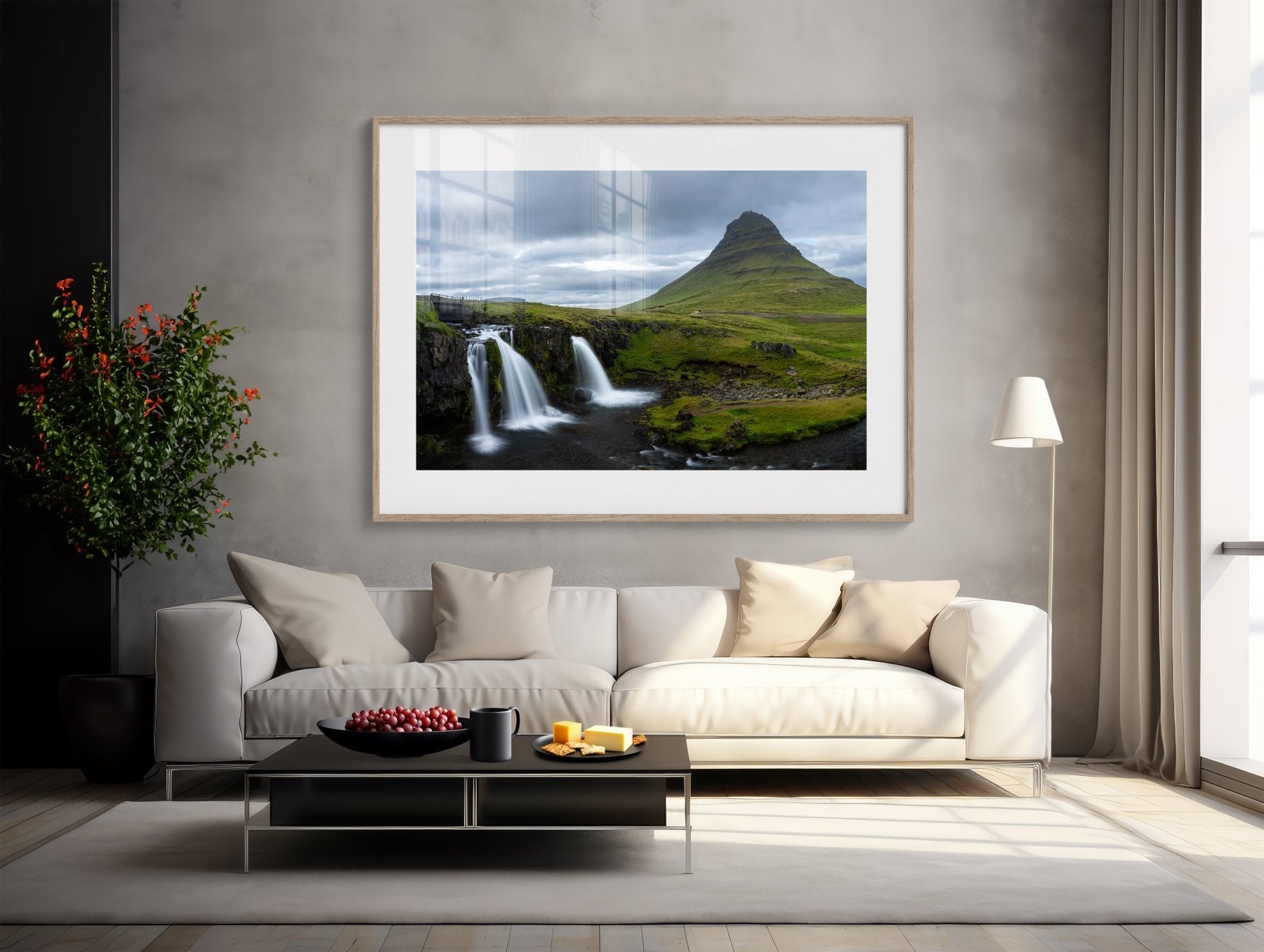 A modern living room with a white sofa, black coffee table, tall floor lamp, potted plant, and a large framed “Kirkjufell Iceland Photography Print – Three Waterfalls One Mountain” on a gray wall.
