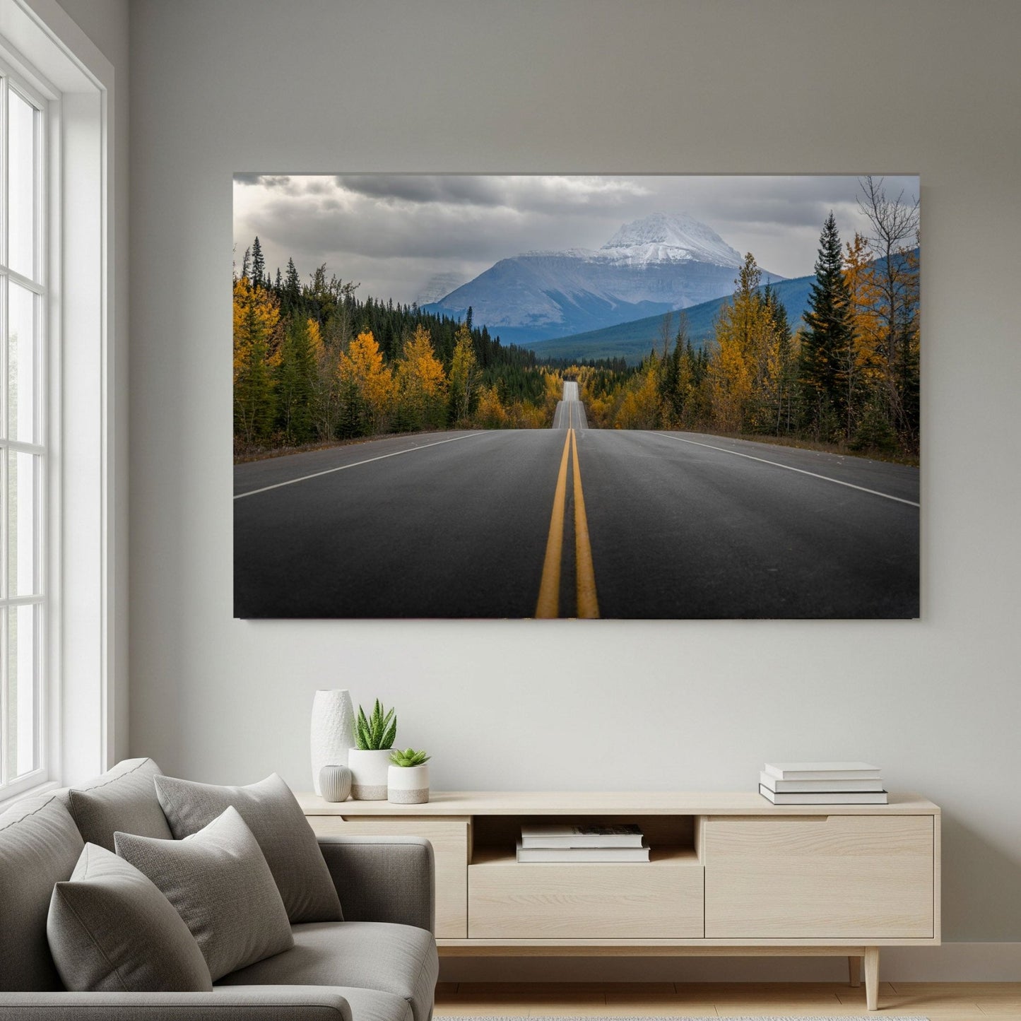 A living room with a gray sofa and light wood furniture features "Road to Autumn | Icefields Parkway, Canadian Rockies Print," showing an empty road through autumn trees leading to a snow-capped mountain under a cloudy sky in Banff National Park.