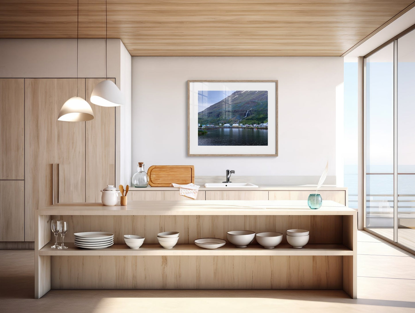 A modern, minimalist kitchen with light wood cabinets, pendant lights, and a large window overlooking the sea features “The Perfect Pair - Iceland Photography Print | Seyðisfjörður Harbor Houses” above the sink.