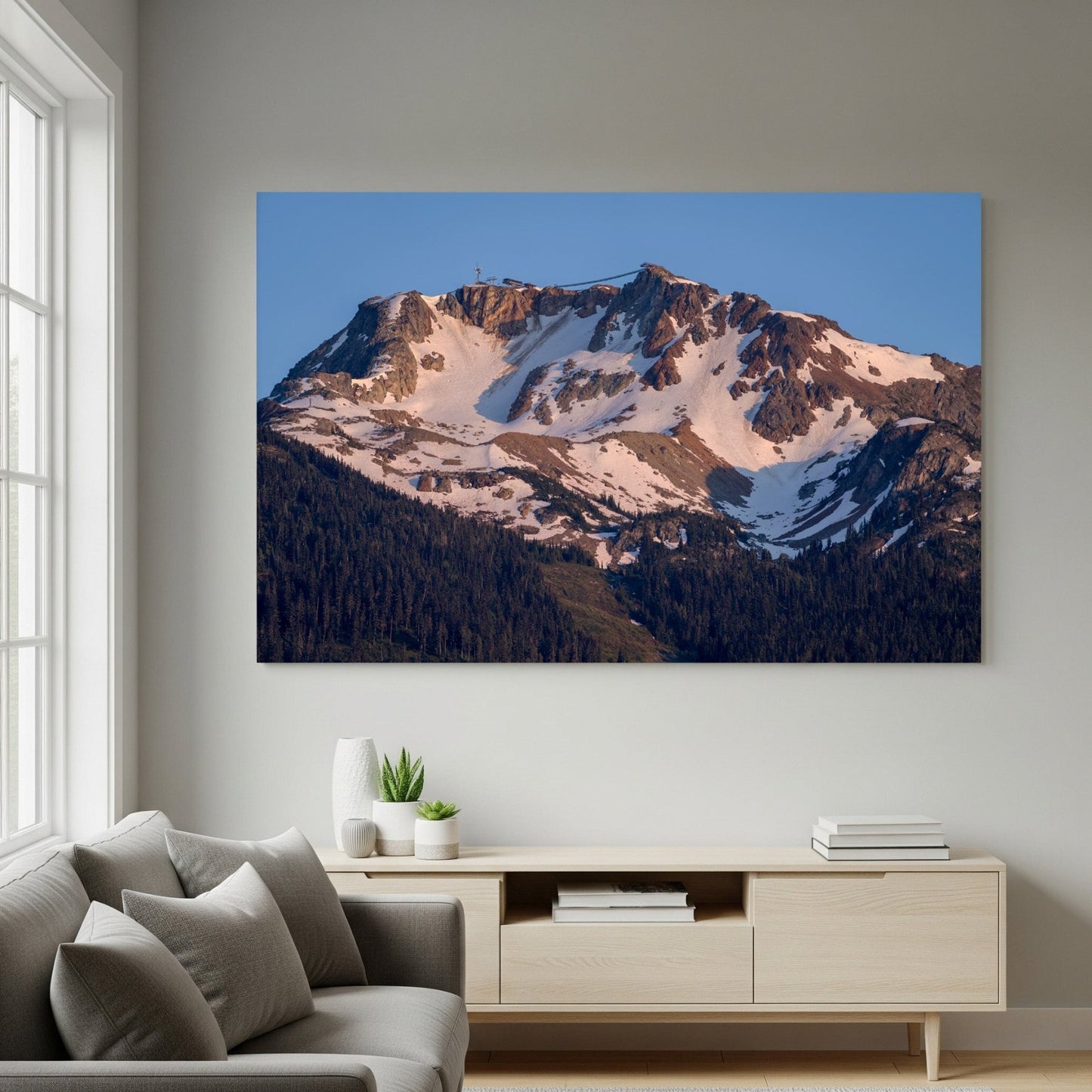 A modern living room features a gray sofa, cushions, and a light wooden console table. Above it hangs "Whistler Alpine Glow | Canadian Mountain Wall Art," capturing Whistler Mountain at sunset. Natural light streams through the window.