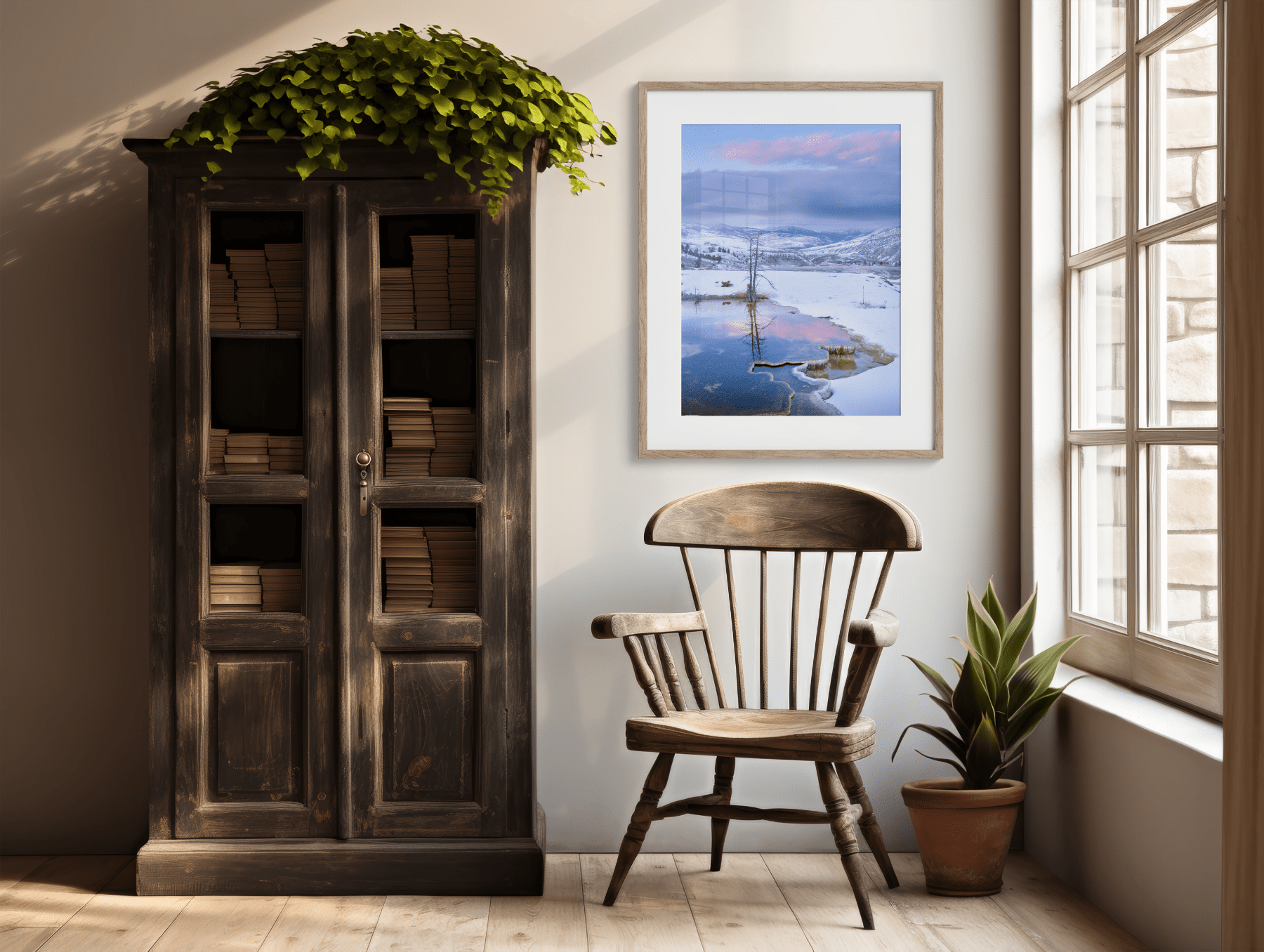 A cozy reading nook with a wooden chair, potted plant, tall vintage bookcase brimming with books, and "Winter's Water Color - Mammoth Hot Springs Yellowstone Photography Print," all bathed in natural sunlight from a large window.